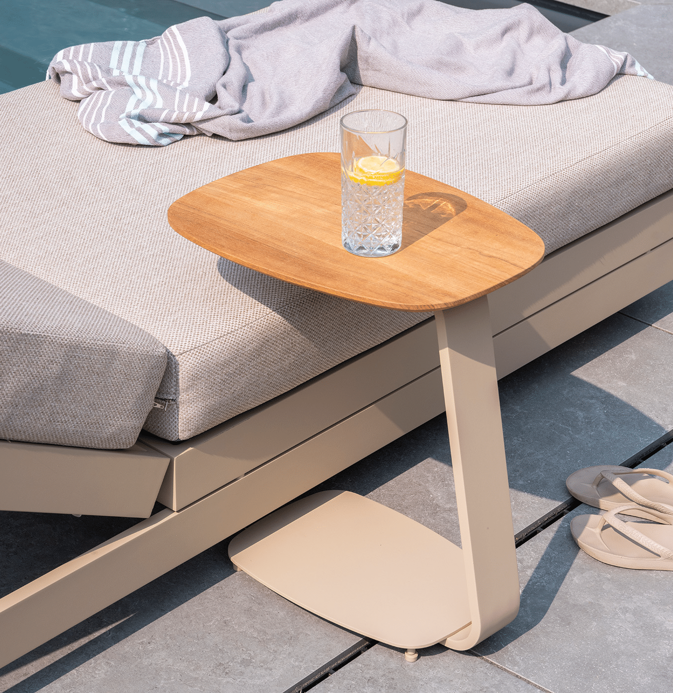 Gentle side table with teak finish in lifestyle environment next to a sunlounger in garden.